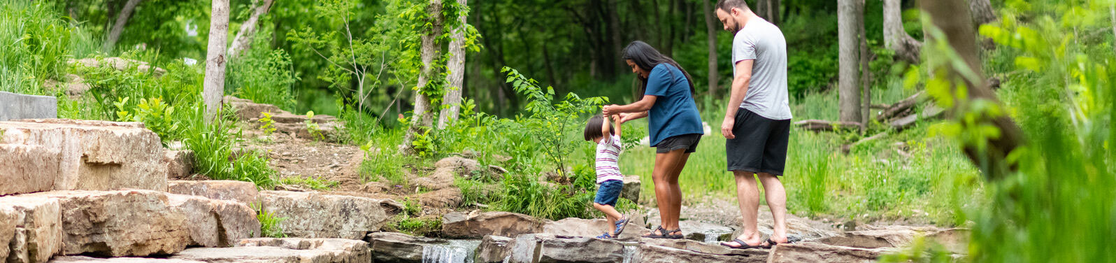 A small child walks across stepping stones in a creek. His mom holds both his little hands from above while his dad walks behind.