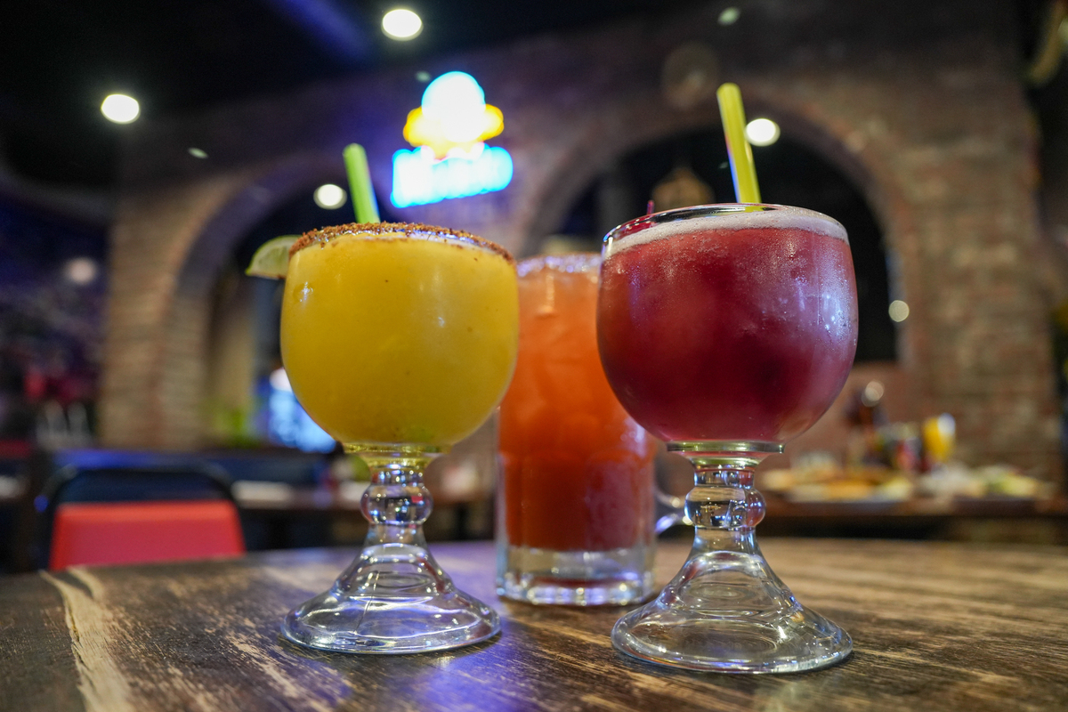 Three colorful margaritas on a table with neon sign behind it. 