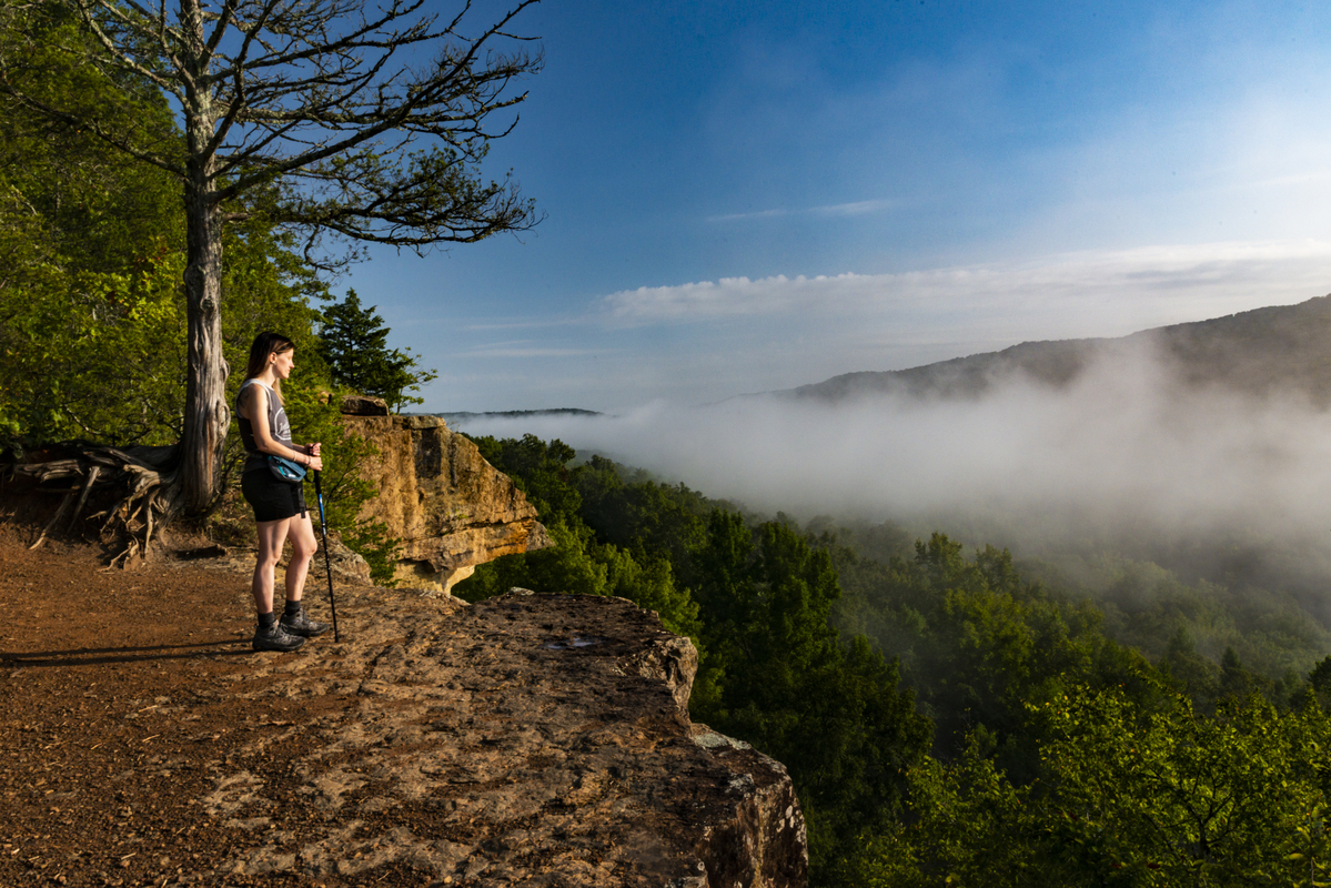 Female with walking stick in hand standing on top of a rock bluff overlooking the wilderness below. 